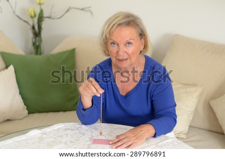 Elderly woman swinging a pendulum over a pack of card in a soothsaying or fortune telling concept as she sits in her living room relaxing