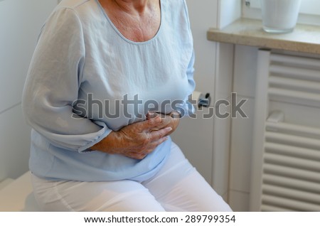 Mid section of senior woman wearing white trousers and light blue blouse sitting on toilet holding stomach