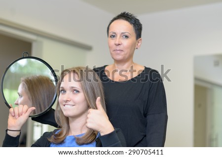Pretty smiling 20s woman with light brown hair wearing blue top and black cardigan gives thumbs up sign, female hairdresser with short dark hair in black top stands behind holding mirror smiling