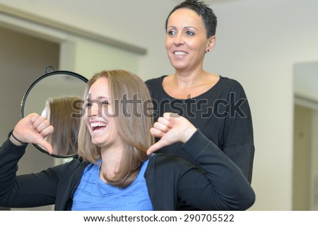 Laughing 20s woman with light brown hair wearing blue top and black cardigan giving thumbs down sign to smiling female hairdresser with short dark hair in black top showing her new haircut in mirror