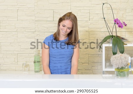 Waist up front view of elegant young 20s woman with light brown hair standing behind white counter in spa or beauty salon wearing blue top smiling and looking down, purple orchid plant in foreground