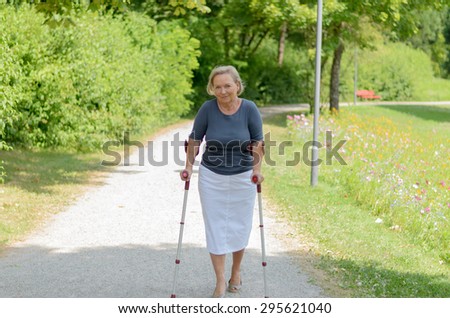 Elderly lady walking in the country along a rural lane on crutches as she tries to exercise and stay fit despite a handicap or injury