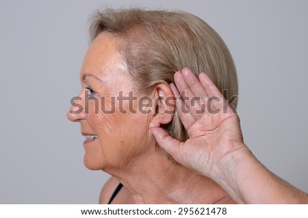 Elderly lady with hearing problems due to ageing holding her hand to her ear as she struggles to hear, profile view on a grey background