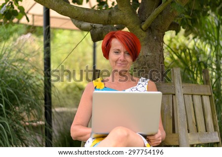 Adult Redhead Woman with Laptop Computer, Sitting on the Wooden Bench Under the Tree and Smiling