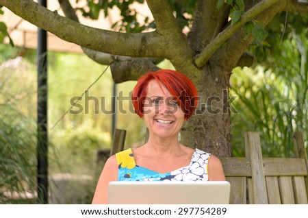 Adult Redhead Woman with Laptop Computer, Sitting on the Wooden Bench Under the Tree and Smiling at the Camera