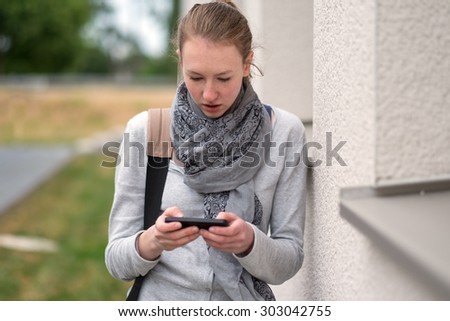 Serious attractive trendy young woman in a scarf standing outside a rural building holding a mobile phone while texting