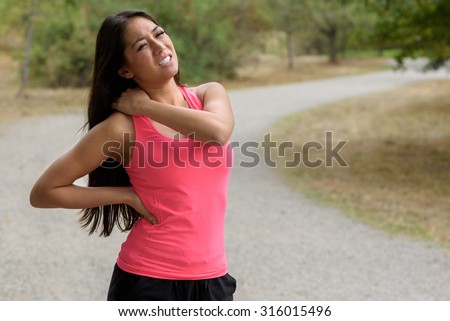 Young woman out jogging suffers a muscle injury standing holding her neck and lower back while grimacing in pain on a rural road, close up upper body view