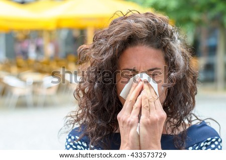 Young woman with lovely long curly hair standing outdoors blowing her nose in an urban square due to a seasonal cold or hay fever
