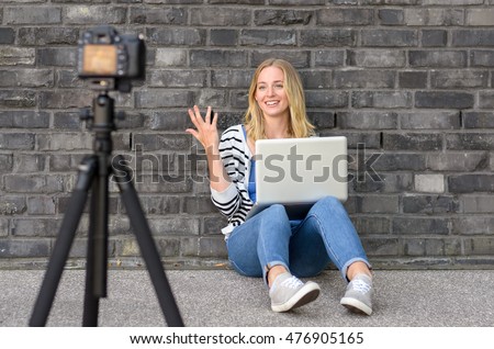 Cute blond female blogger with laptop recording video while sitting and talking with gesturing hands for theme about video blogging