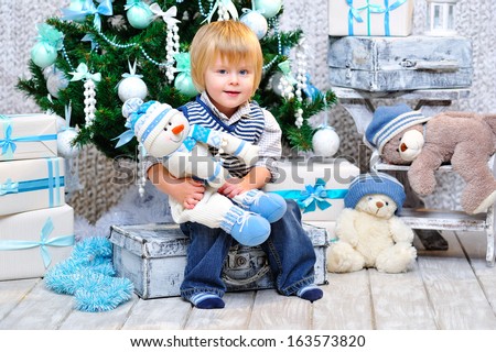 Cute little boy sitting down by the decorated Christmas tree with toys, teddy bears and gift boxes