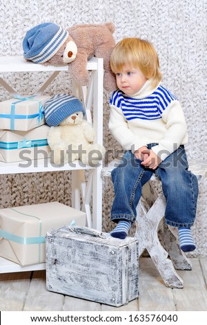 Cute little kid sitting on a white wooden chair next to the shelf with teddy bears and gift boxes and dreaming about Christmas