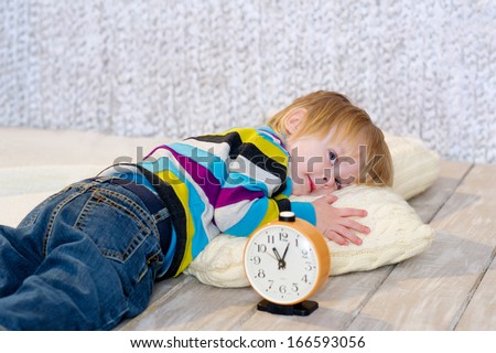 Cute toddler trying to fall asleep lying down on the wooden floor with clock