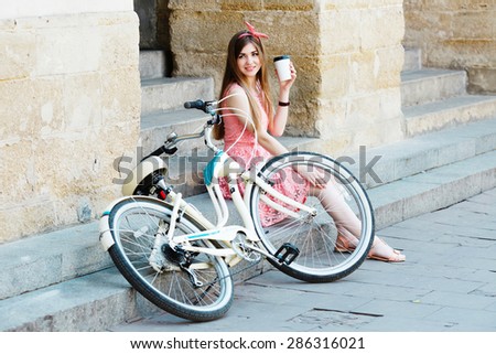 Young pretty blond-brown girl sitting on concrete stairs with a cup of coffee and looking toward, with vintage bicycle, in an old European city