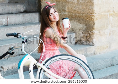 Young pretty blond-brown girl sitting on concrete stairs and smiling with a cup of coffee, looking at camera, with vintage bicycle, in an old European city
