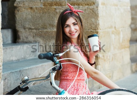 Young pretty blond-brown girl smiling with a cup of coffee and vintage bicycle, looking at camera