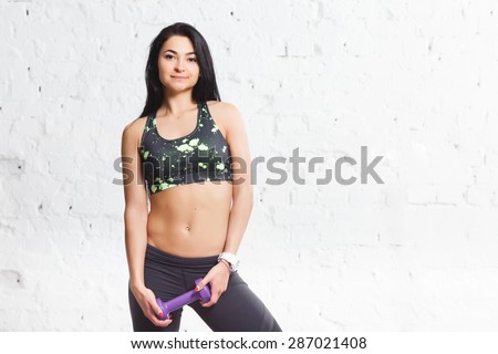 Athletic young brunette woman smiling and holding purple dumbbells, with strong abs and flat belly, in sports outfits with beautiful body, against concrete wall. Copy space