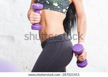 Close up athletic young woman showing some strong abs and flat belly, in sports outfits with beautiful body, holding purple dumbbells, against concrete wall