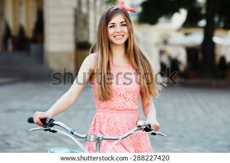 Pretty blond-brown girl smiling and holding bicycle. Wearing pink head wrap and dress with a pattern of flowers. Looking at the camera. In the old European city. Close up.
