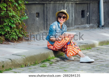 Happy smiling woman tourist sitting on the curb with tablet. In European city.