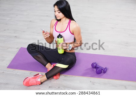 Beautiful brunette young girl with smart phone sitting on yoga mats, workout break. Texting and social networking.