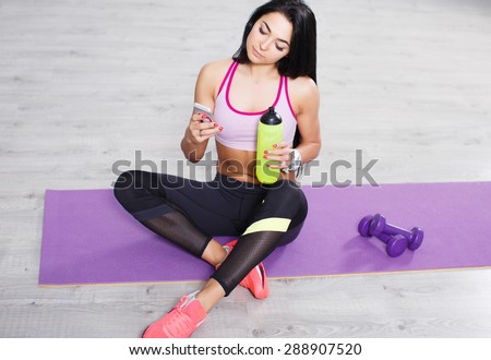 Beautiful brunette young girl with smart phone sitting on yoga mats, workout break. Social networking and holding a sports drink.