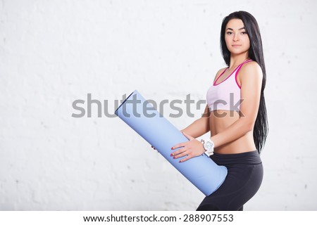 Close-up portrait of young pretty fitness brunette girl in sports wear holding blue yoga mat over wall background