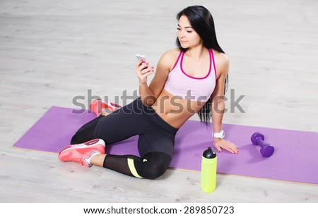 Beautiful brunette young woman with smart phone sitting on yoga mats, workout break. Texting and social networking.