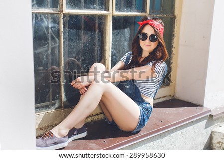 Lifestyle urban portrait of pretty brunette girl posing at denim overall bright bandana and sunglasses. Sitting on the windowsill outdoor.