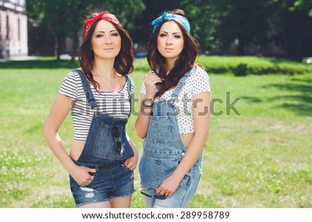 Outdoor lifestyle portrait of two twins sisters best friends hipster girls wearing stylish bright outfits, bandanas, denim overalls, having great time together. Against of park background.