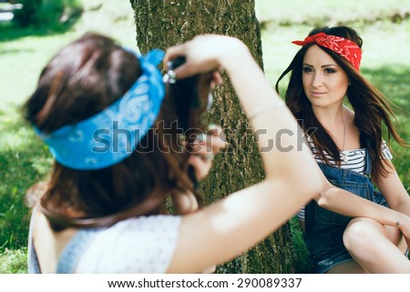 Close up of brunette girl in blue bandana holding digital camera and making photo of pretty brunette girl in red bandana on the park. Summer holidays.