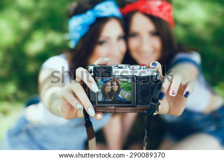 Portrait of twins girl on the screen of digital camera. Girls taking selfie with camera in the park. Wearing colorful bandanas and denim jumpsuits.