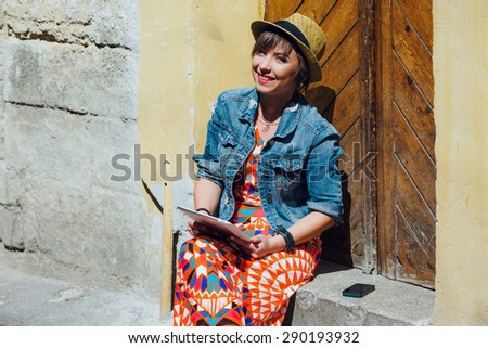 Colorful trendy tourist girl smiling and sitting on the curb with tablet, against the background of wooden doors. Looking at camera.