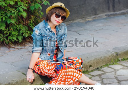 Young pretty woman colorful wearing sunglasses and hat. Sitting on the curb with tablet and looking at the camera. Copy space.
