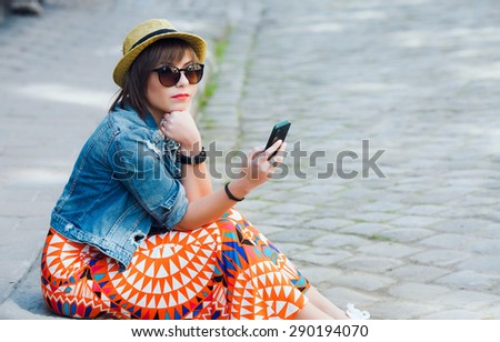 Trendy cool hipster woman colorful wearing sunglasses and hat. Sitting on the curb and looking at the mobile phone. Copy space.