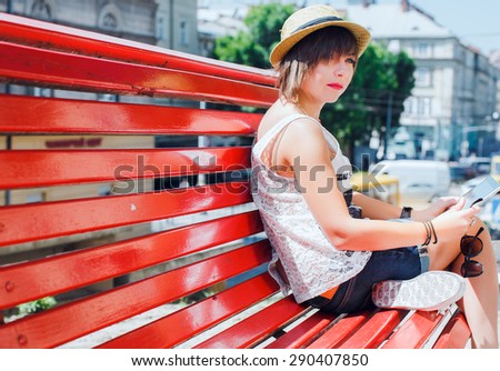 Cute girl with shortcut dark-blonde hair. Wearing stylish summer hat, white top and denim shorts. Sitting on red bench and holding tablet, on city background.
