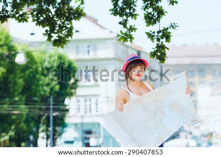 Pretty tourist girl holding a big map. On city background. With shortcut dark-blonde hair. Wearing stylish pink hat and denim shorts. Looking at camera.