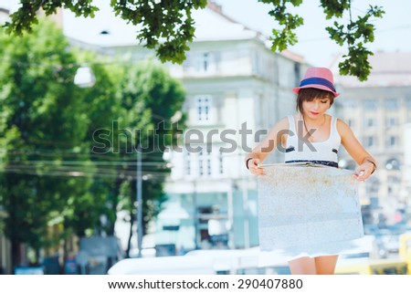 Pretty tourist girl looking at map. On city background. With shortcut dark-blonde hair. Wearing stylish pink hat and denim shorts. Looking at camera.