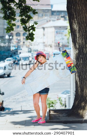 Cute tourist young woman speaking on the mobile phone and holding a big map of Europe. On city background. With shortcut dark-blonde hair. Wearing stylish pink hat and denim shorts.