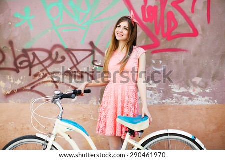 Pretty young girl smiling with long blond-brown hair, in pink head wrap and dress with a pattern of flowers. Holding vintage bicycle on urban graffiti background.