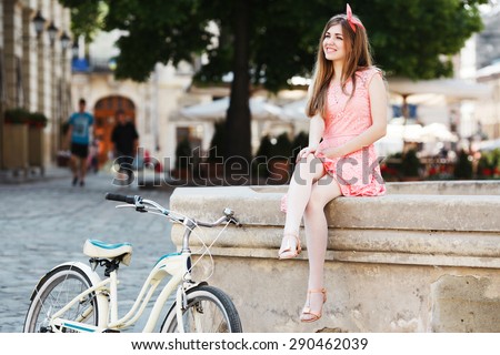 Happy blond-brown girl smiling and sitting on a fountain. Wearing pink head wrap and dress with a pattern of flowers. With retro bicycle nearby. In the old European city.