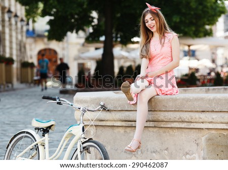 Funny blond-brown girl having fun with her feet. Wearing pink head wrap and dress with a pattern of flowers. With retro bicycle nearby. In the old European city.