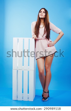 Full-length portrait of brunette attractive young woman in pink dress, leaning on the white box. Studio portrait. On blue background.