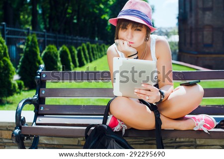 Young thoughtful tourist woman sitting on bench with digital tablet. In bright pink hat. On city background.