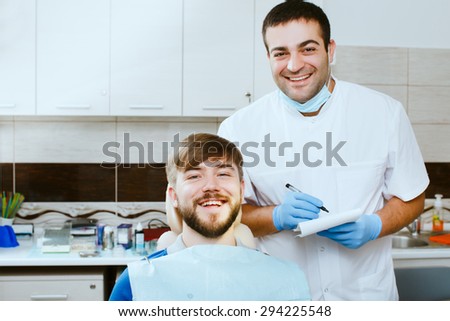 Happy male dentist taking notes and looking at camera, with male patient smiling, at the dental office.