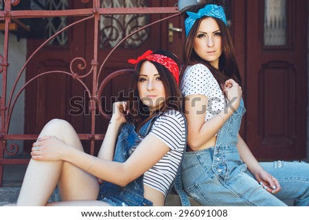 Portrait of pretty young brunette twins girls. Wearing denim overalls, bright bandanas and white top, posing outdoors. On old doors background. Looking at camera. Summer day.