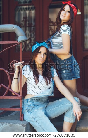 Portrait of beautiful young brunette twins girls, on concrete stairs. Wearing denim overalls bright bandanas, posing outdoors. On old doors background. Looking at camera. Summer day.