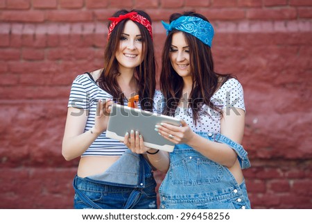 Portrait of pretty brunette twins girls with tablet and smart phone on brick wall background. With long dark-brown straight hair. Wearing jeans overalls, red and blue bandanas and white top.