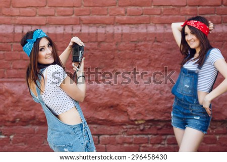 Brunette twins woman. Girl in blue bandana holding digital camera and taking photo of pretty brunette girl in red bandana posing on the brick wall background. Both wearing denim overalls and white top