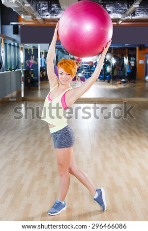 Young redhead woman with short hair is standing near the mirror with fitball in gym, full length
