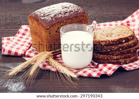 Freshly baked loaf of bread with spikelets of wheat and glass of milk on a red and white cloth on a dark wooden boards. close up
